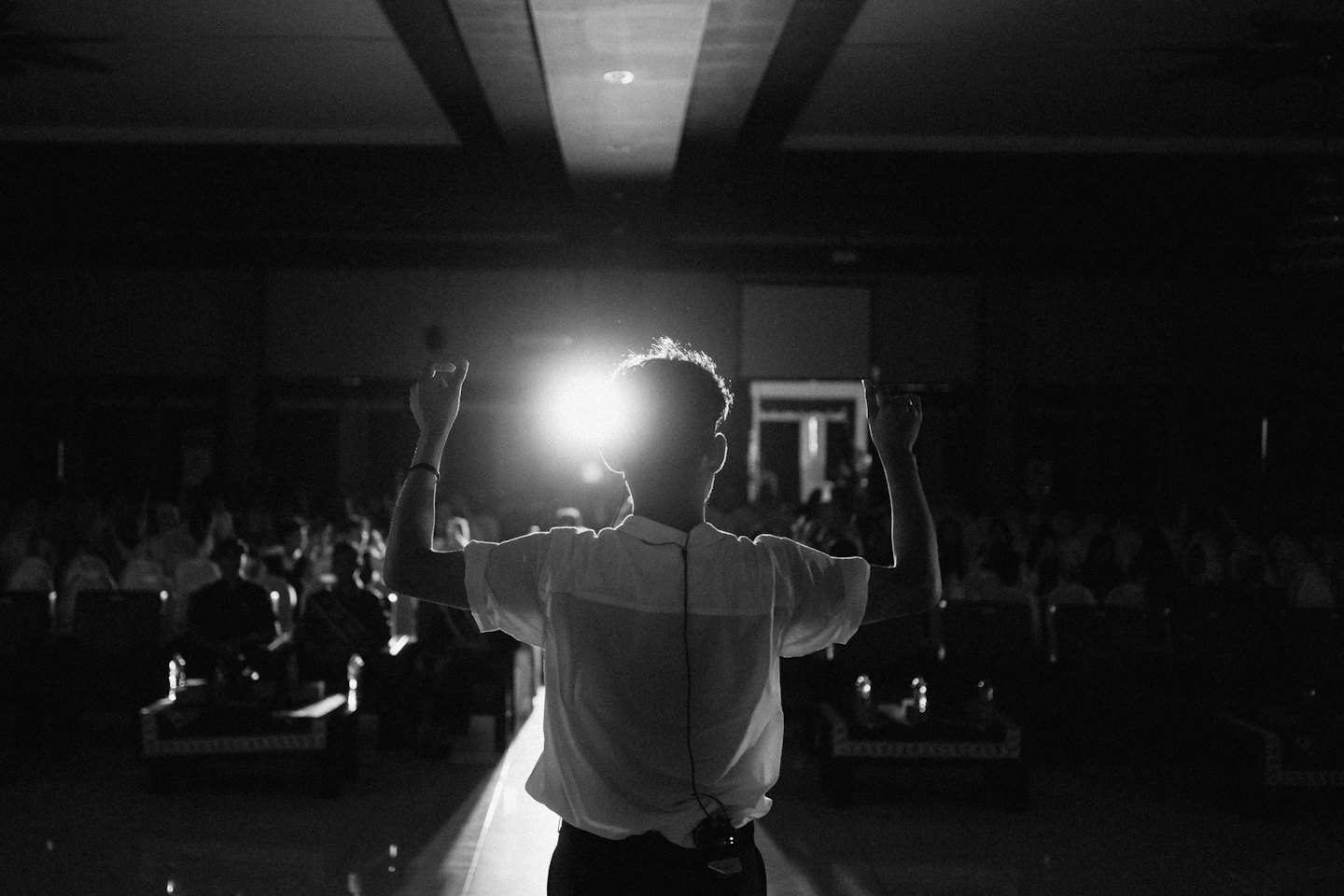 Speaker stands on stage with arms raised before an audience.